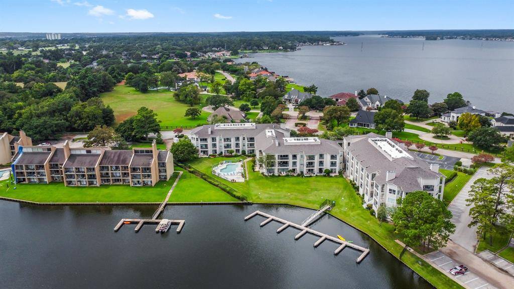 waterfront property with multiple residential buildings, each featuring balconies overlooking a serene lake. The area includes well-kept lawns, a swimming pool, and docks for water access. The surrounding neighborhood is lush with greenery, offering a peaceful, scenic environment.