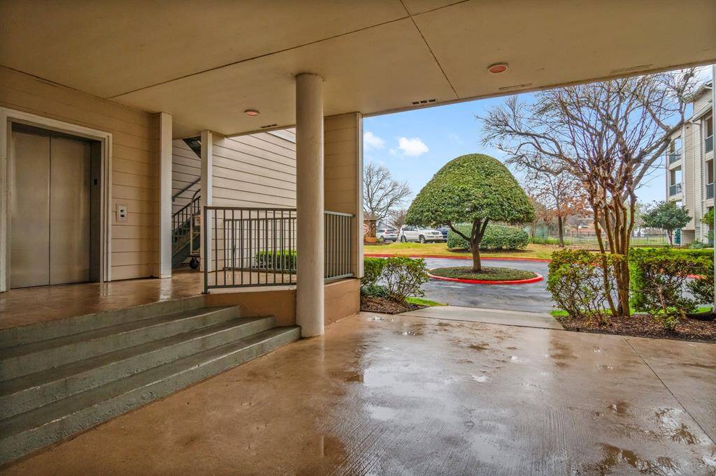 a covered entrance area of a residential building with an elevator and stairs. It opens to a landscaped view, featuring a neatly trimmed tree and a driveway. The space appears clean and well-maintained.