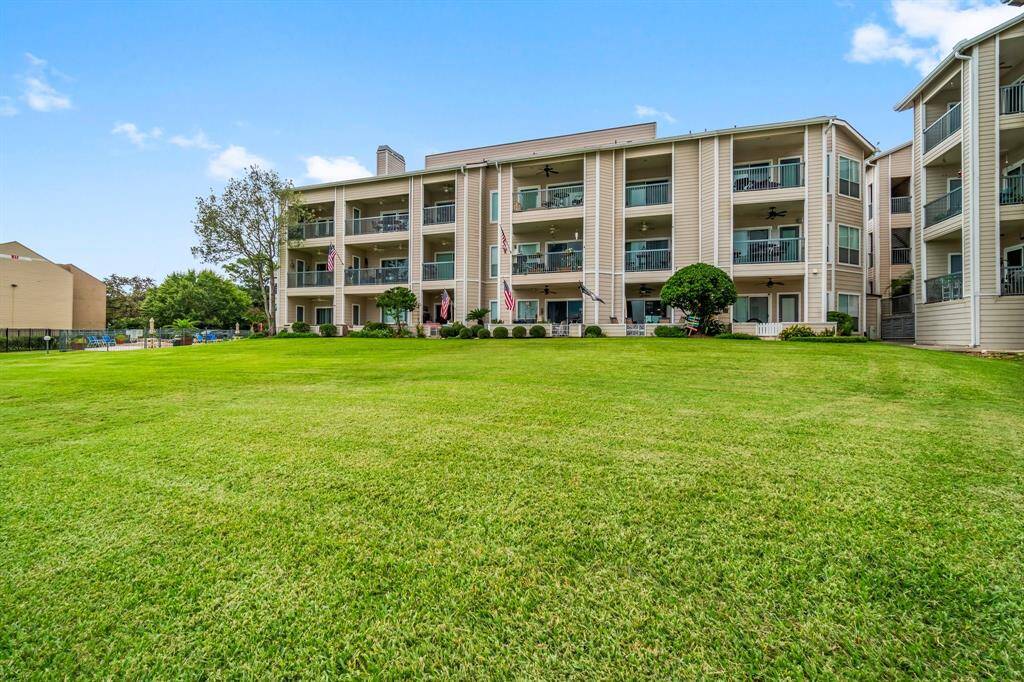 a well-maintained multi-level complex with spacious balconies, set against a large, lush green lawn. The building features modern siding and is surrounded by trees and shrubs, providing a serene environment.