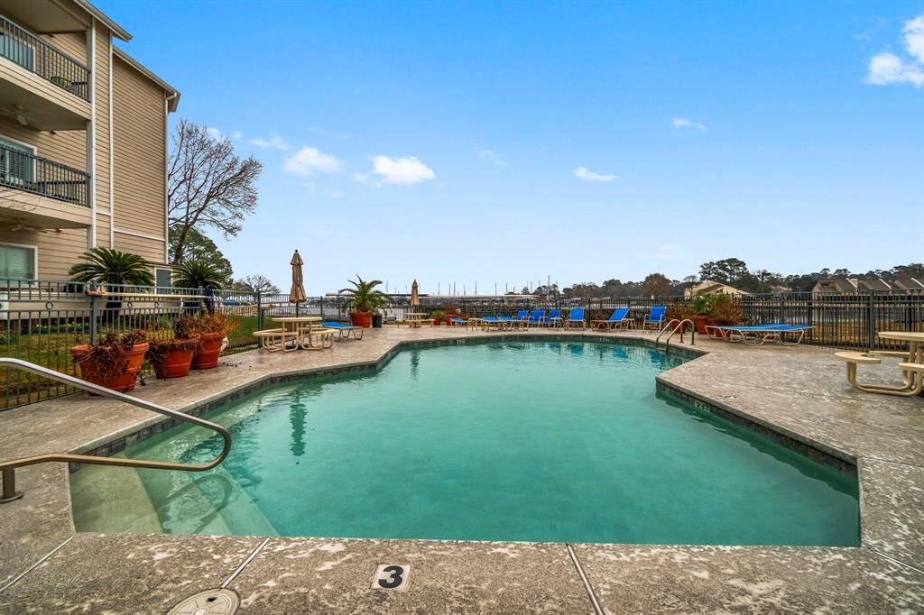 serene outdoor pool area with lounge chairs and tables, surrounded by a secure fence. The pool is adjacent to a multi-story residential building, offering a relaxing setting with views of a marina and greenery in the distance.