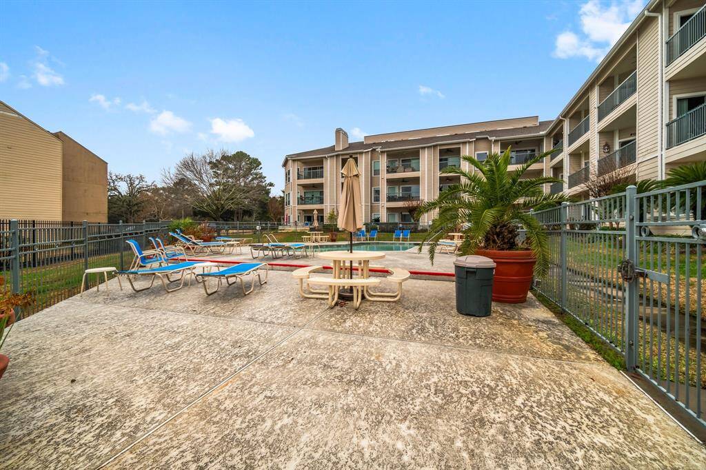 Spacious outdoor area featuring a fenced patio with sun loungers and a table, surrounded by landscaped greenery, adjacent to a multi-story residential building. Perfect for relaxation and outdoor activities.