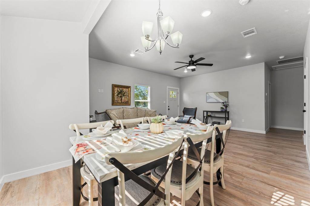 This photo showcases a bright, open-concept living and dining area with light wood flooring. It features a modern chandelier above a dining table set for six and a cozy seating area with a ceiling fan. The room has neutral walls, ample natural light, and tasteful decor, creating a welcoming atmosphere.