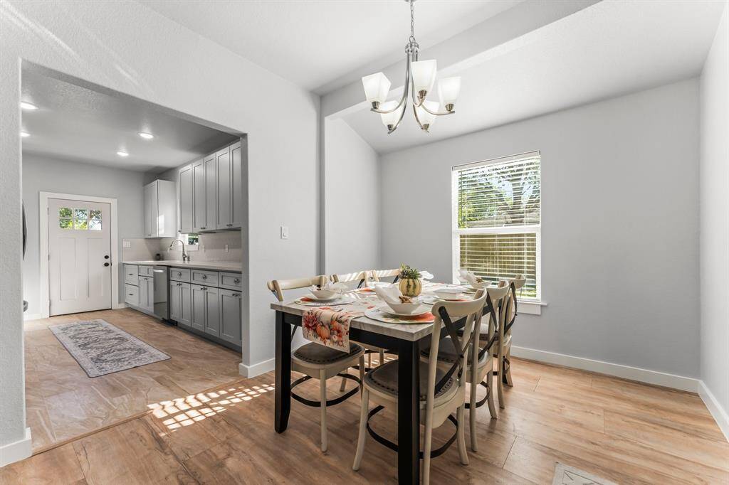 This photo showcases a bright dining area with a stylish chandelier and a window letting in natural light. Adjacent to it is a modern kitchen featuring gray cabinets, ample counter space, and a light-colored tiled floor, creating a welcoming and functional space.