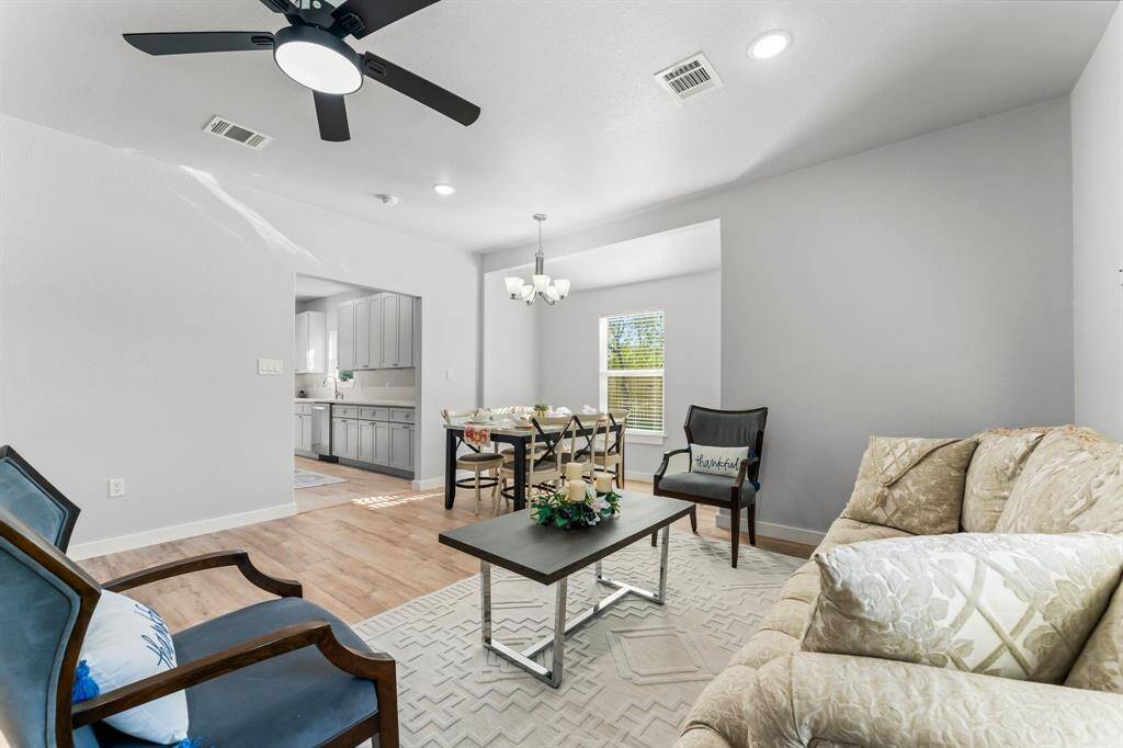 This photo showcases a bright, open living and dining area with modern furnishings. The room features light-colored walls and wood flooring, with a ceiling fan and recessed lighting. Adjacent is a view into a kitchen with sleek cabinetry.