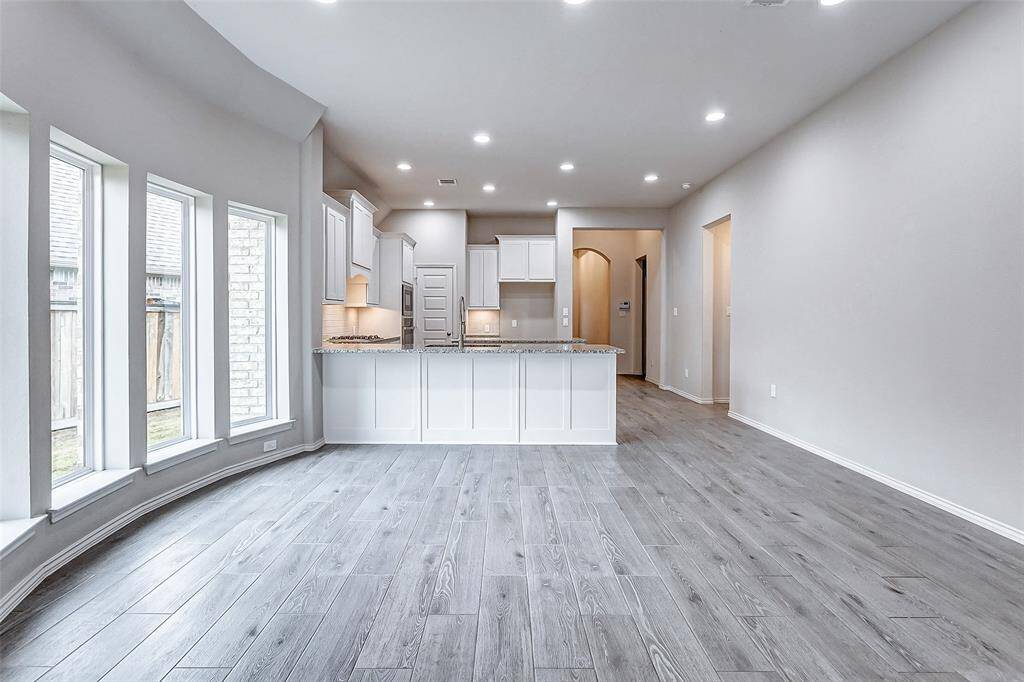 This photo showcases a bright, open-plan kitchen and living area with modern finishes. The kitchen features a large island, white cabinetry, and recessed lighting. Wide windows provide natural light, complementing the sleek gray flooring. The space is inviting and ideal for entertaining.