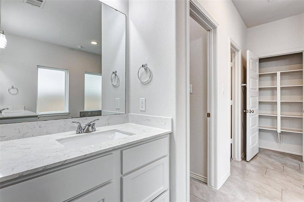 Modern bathroom with a sleek marble countertop and sink. It features a large mirror, frosted windows for privacy, and an adjacent area with built-in shelving, offering ample storage space. The light-color tiles and walls create a bright, clean atmosphere.