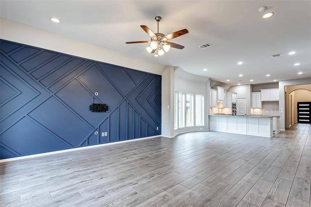 This spacious open-plan living area features modern wood-like flooring, a striking navy accent wall, and ample natural light. The room flows seamlessly into a bright kitchen with white cabinetry and a large island, perfect for entertaining.