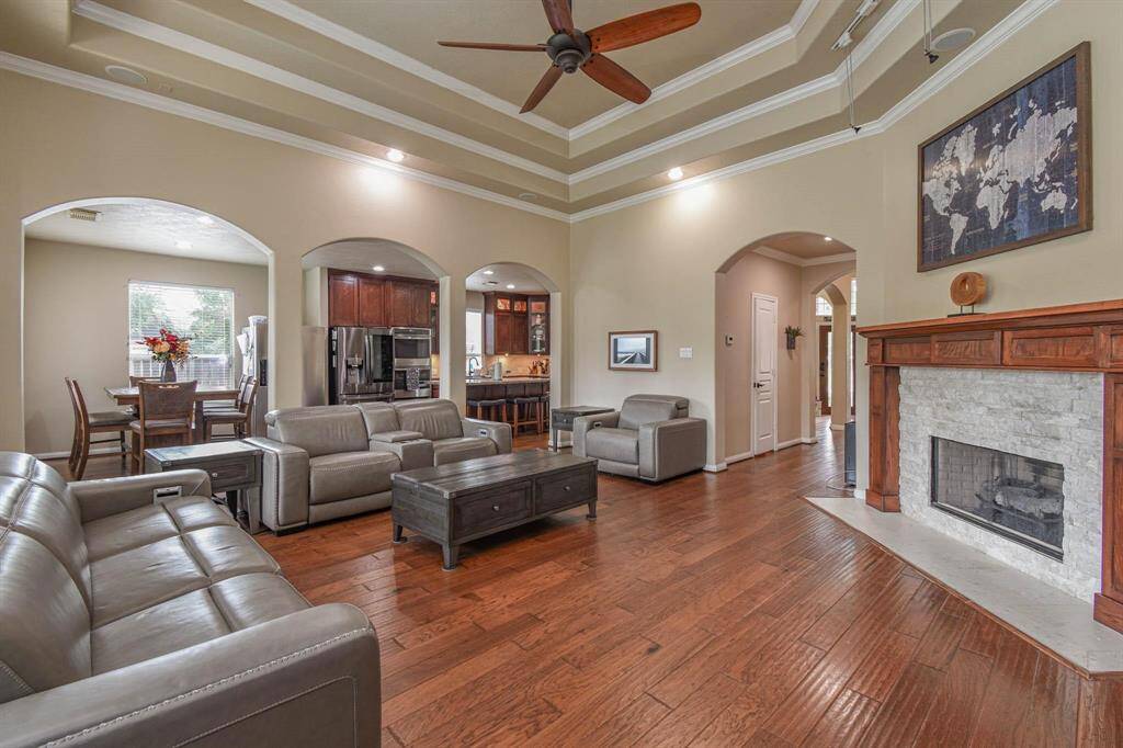 Spacious living room with hardwood floors, featuring a cozy fireplace and elegant tray ceiling with recessed lighting. The open layout connects to a modern kitchen and dining area, creating a welcoming atmosphere for entertaining.