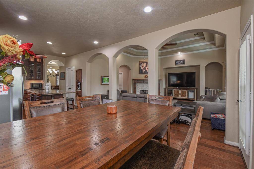 This photo showcases an open-concept living area with a dining table in the foreground. The space features archways, recessed lighting, and wood flooring. A modern kitchen with stainless steel appliances is visible, along with a cozy living room that includes a fireplace and large TV.
