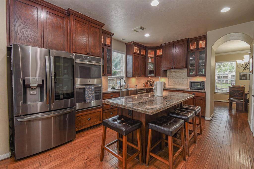 This kitchen features rich wooden cabinetry, a large granite island with seating, stainless steel appliances, and hardwood floors. It's well-lit with recessed lighting and has a cozy dining area visible through an arched doorway.