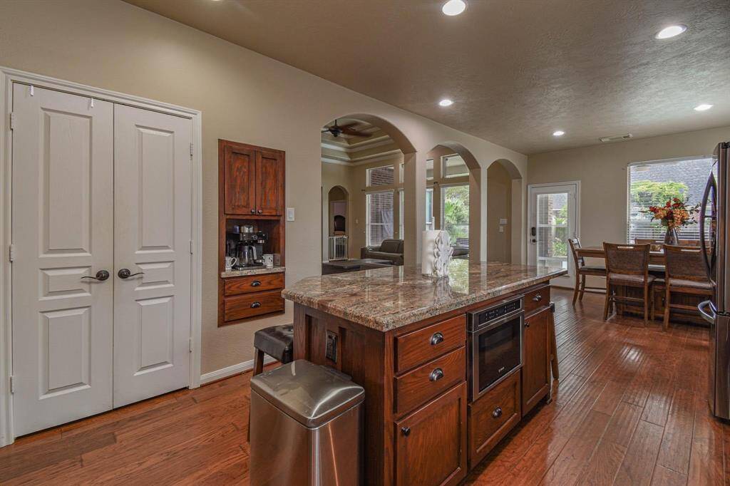 This photo showcases a spacious, open-concept kitchen and dining area with modern lighting, a large granite island, and hardwood floors. It features a built-in coffee station, stainless steel appliances, and ample natural light through large windows and glass doors, leading to a cozy living space.