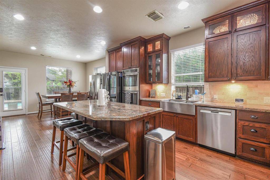 This kitchen features rich wooden cabinetry, granite countertops, and stainless steel appliances. It includes a large island with seating and a farmhouse sink. The space is bright with natural light from multiple windows, and there's a cozy dining area adjacent to the kitchen.