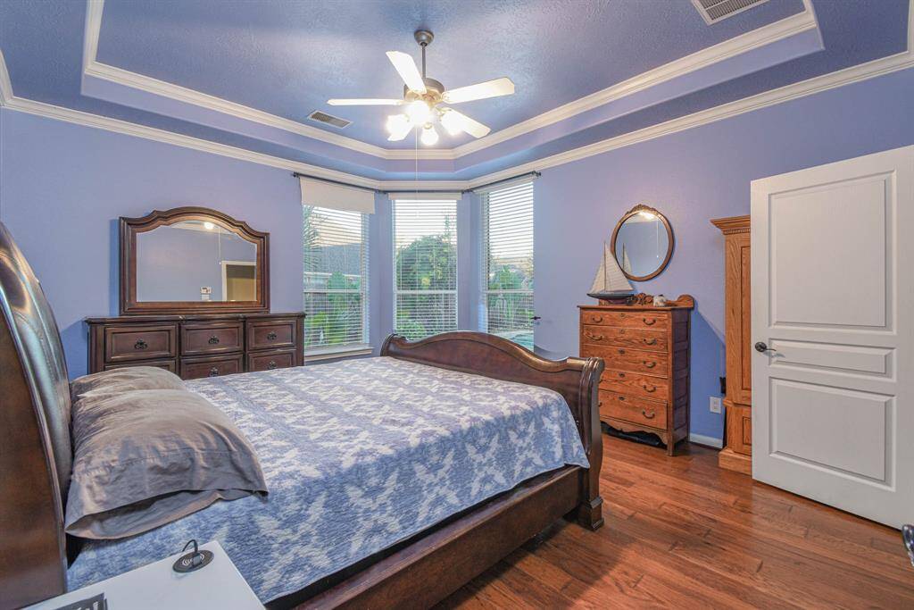 This spacious bedroom features a calming blue palette with elegant crown molding and a tray ceiling. It includes hardwood floors, large windows offering plenty of natural light, and classic wooden furniture that adds warmth and style.