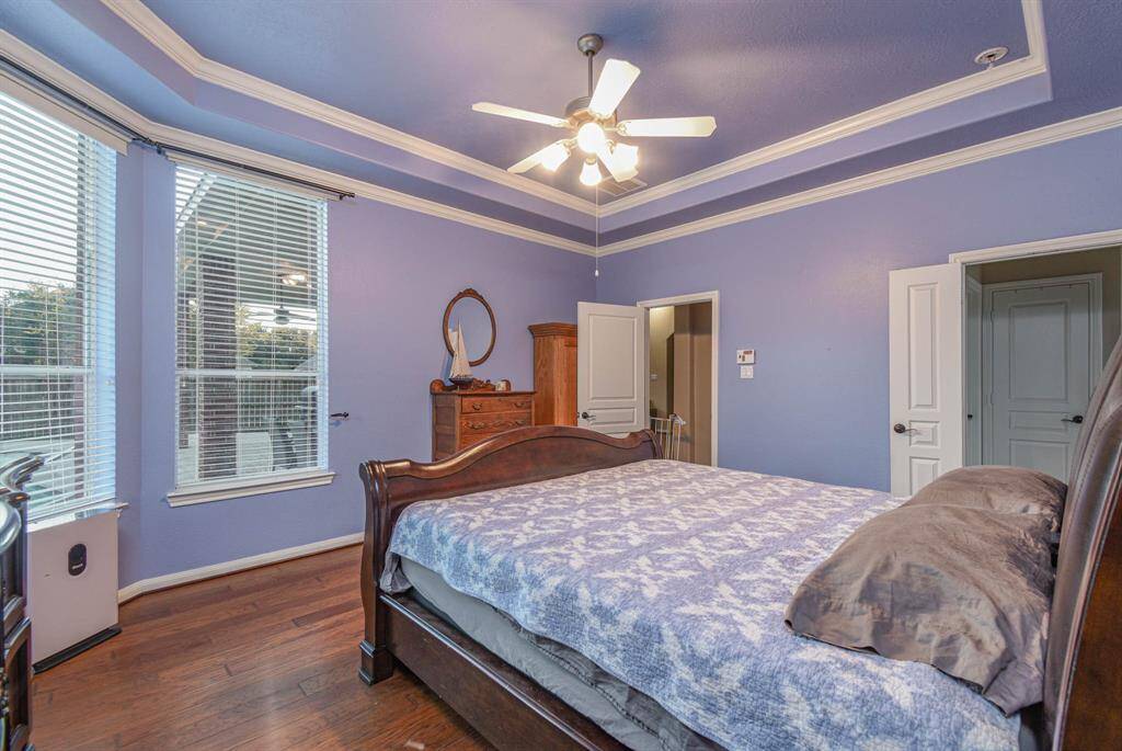 This bedroom features soft blue walls with white trim, a ceiling fan, and large windows that provide ample natural light. It includes wooden flooring and a classic wooden bed, creating a cozy and inviting atmosphere.