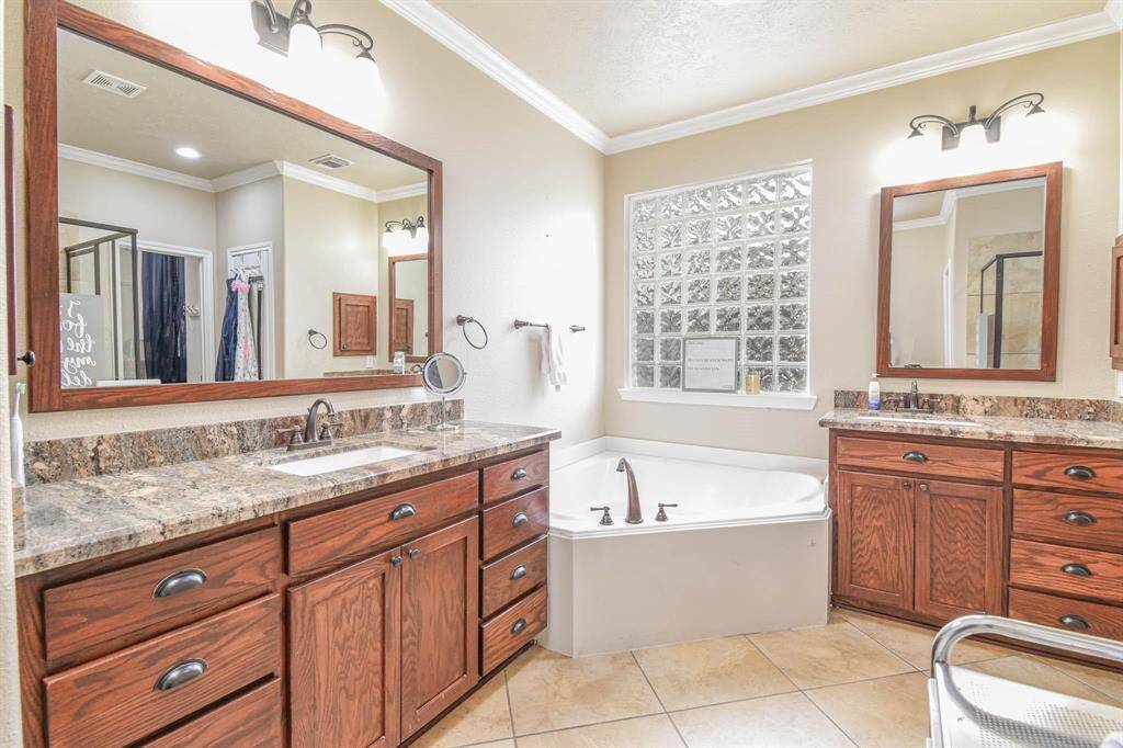 This bathroom features a spacious layout with dual vanities, granite countertops, and rich wood cabinetry. It includes a corner soaking tub, a large mirror, and ample lighting. The tiled floor and frosted glass window add a touch of elegance and privacy.