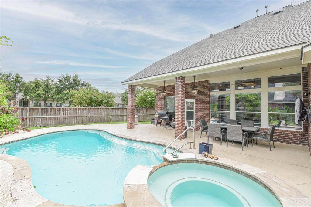 This backyard features a spacious covered patio with ceiling fans, a dining area, and a grill. It overlooks a beautiful pool and spa, surrounded by a privacy fence and landscaped greenery, perfect for outdoor entertainment.