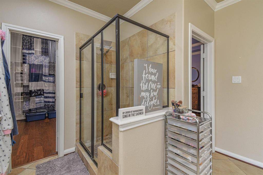 This bathroom features a spacious glass-enclosed shower with neutral tile, a decorative sign, and a nearby metal storage cart. The room connects to Her's and His closets with wood flooring and a curtained doorway, offering a cozy and functional space.
