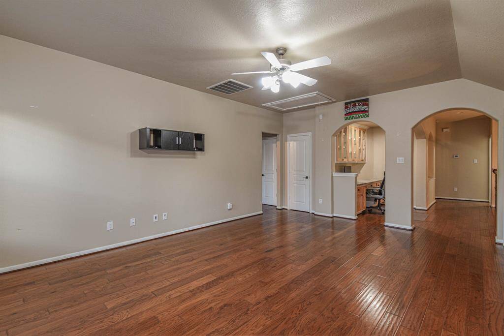 Spacious game room with warm wood flooring and neutral walls. Features a ceiling fan, built-in wall cabinet, and an arched entryway leading to additional rooms. Ideal for a cozy and inviting home atmosphere.