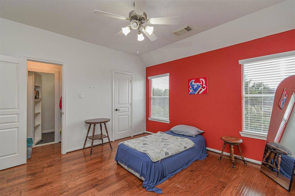 This bedroom features hardwood floors, a bold red accent wall, and ample natural light from two windows. It includes a ceiling fan, closet space, and a simple setup with a bed and side tables.