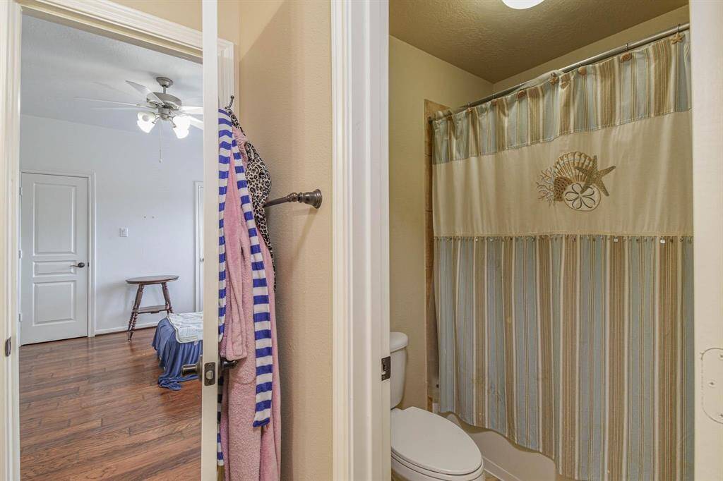 This photo shows a cozy bathroom with a shower featuring a beach-themed curtain. It opens into a bright bedroom with hardwood floors, a ceiling fan, and a simple wooden stool.