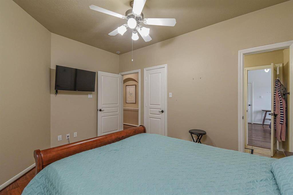 This bedroom features neutral walls, a ceiling fan, and a mounted TV. It includes a wooden bed with a light blue cover, and has two white doors leading to other rooms. The space is well-lit and has a hardwood floor.