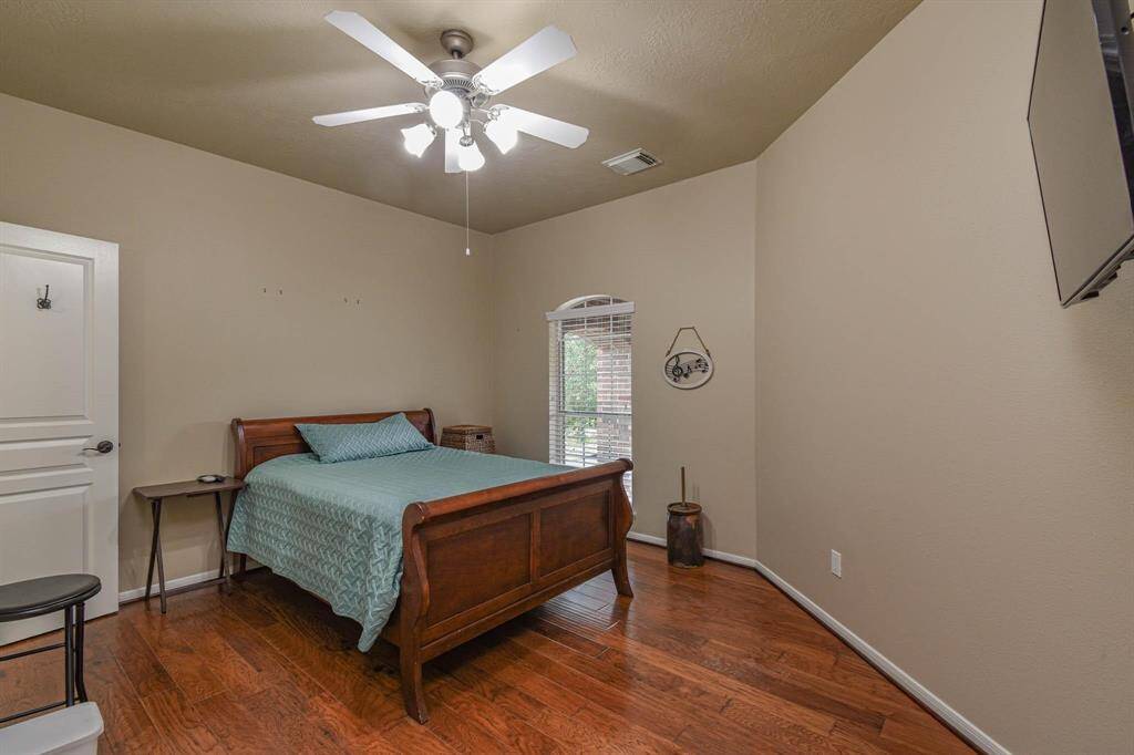 This cozy bedroom features warm wood flooring, neutral walls, and a ceiling fan. It includes a wooden bed, a small side table, and a wall-mounted TV. A window allows natural light to brighten the space.