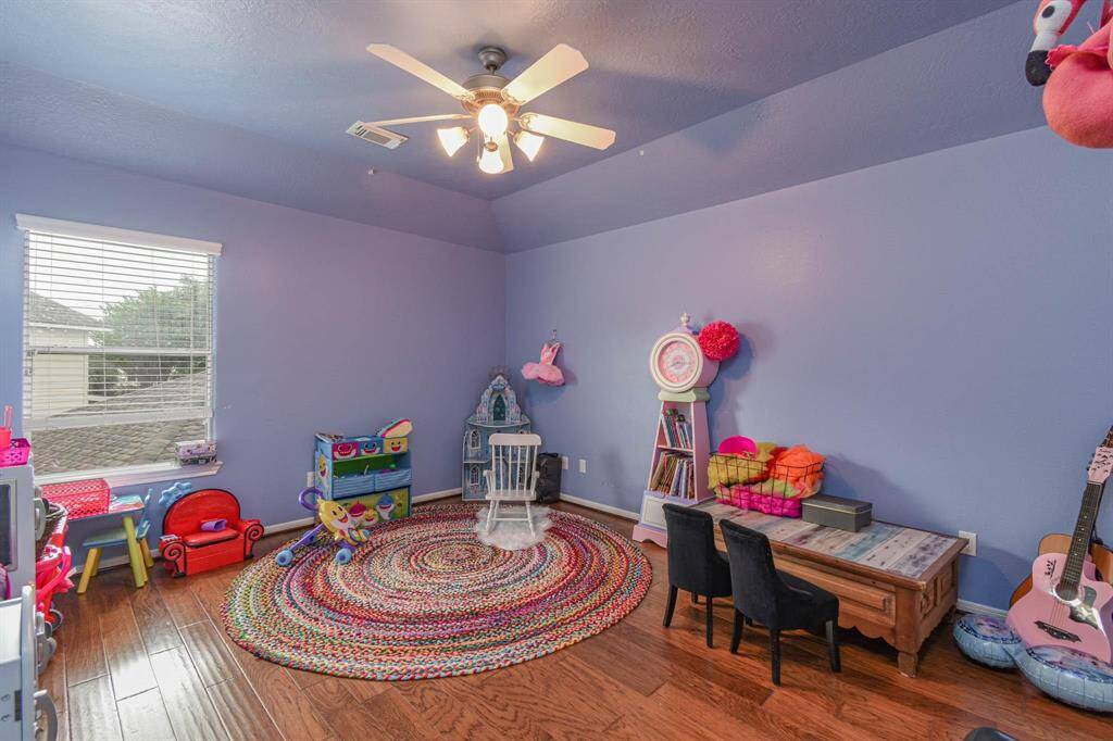 This cozy playroom features blue walls, hardwood floors, and a ceiling fan. It's well-lit with a large window, and includes a colorful circular rug, child-sized furniture, and ample space for toys and activities. Perfect for a child's imaginative play area.