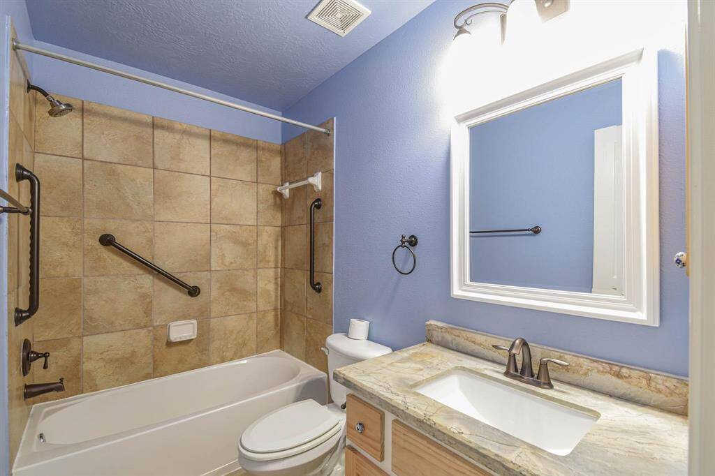 This bathroom features a full-size bathtub with beige tile surround, equipped with safety grab bars. It has a single sink vanity with a marble countertop and a framed mirror above. The walls are painted a soft blue, creating a calming atmosphere.
