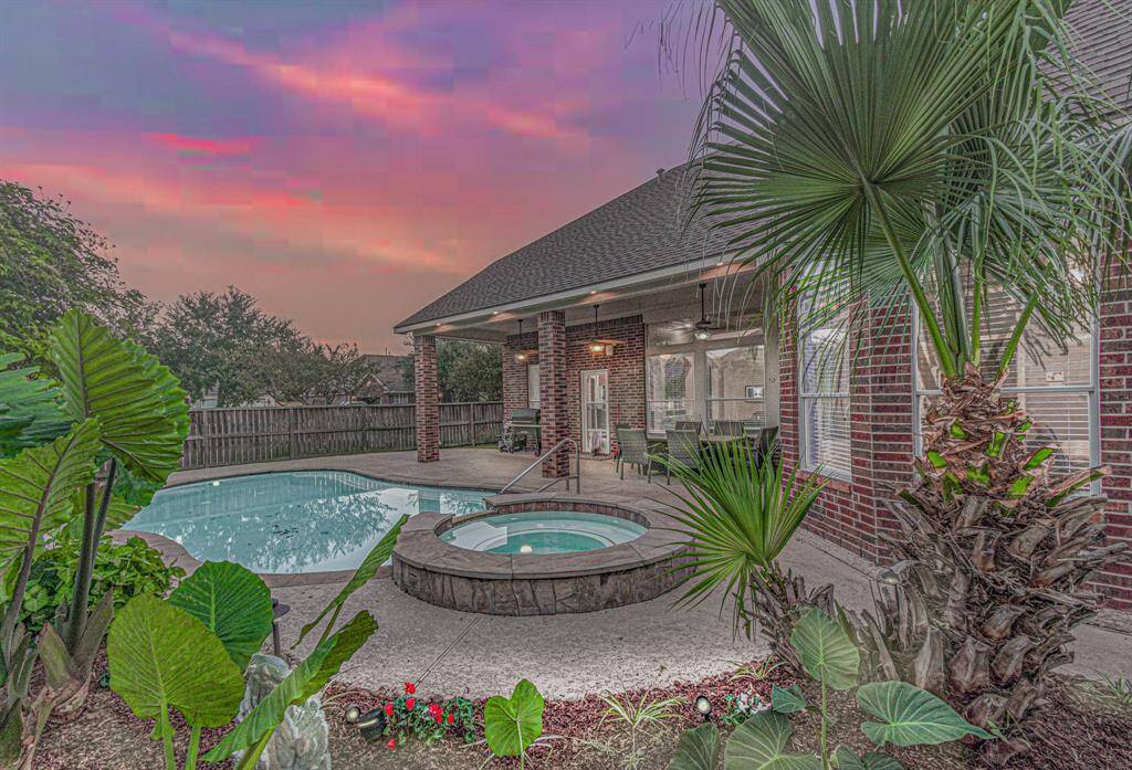 This inviting backyard features a spacious pool and hot tub, surrounded by lush greenery. The covered patio area with outdoor seating is perfect for relaxing or entertaining, all set against a beautiful sunset sky.