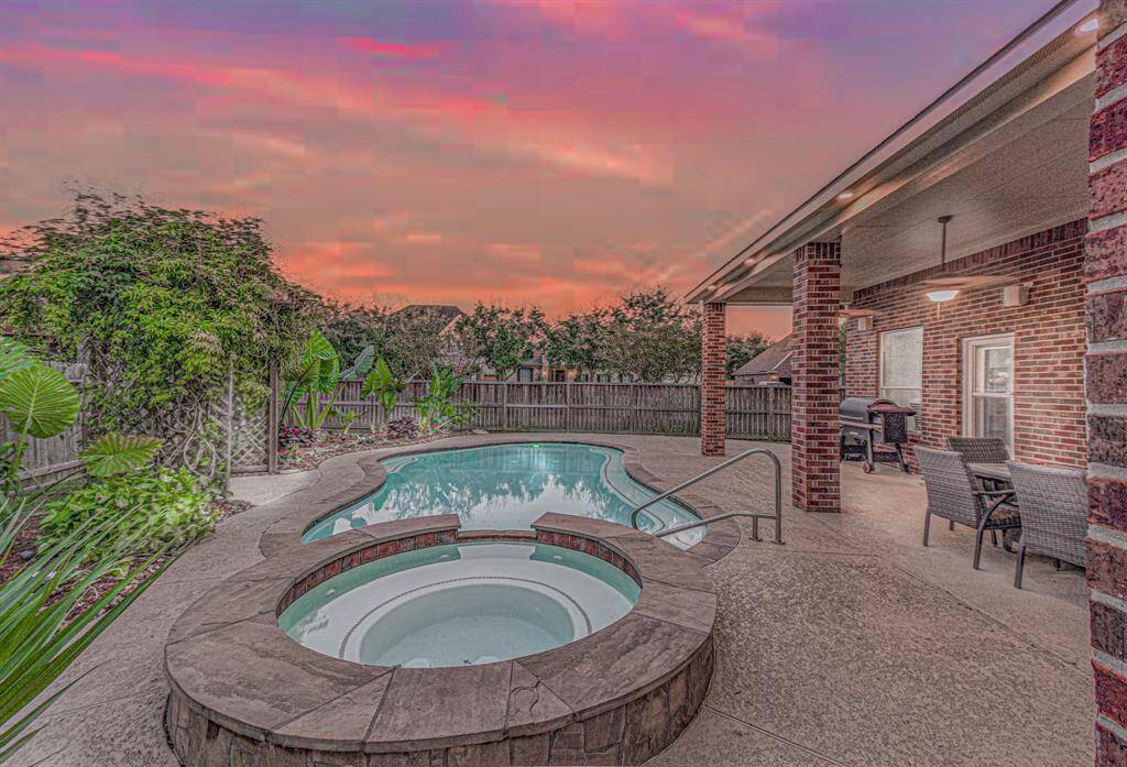This backyard features a swimming pool with an attached hot tub, surrounded by lush greenery and a privacy fence. The covered patio includes seating and a barbecue area, perfect for outdoor entertaining, all set under a beautiful sunset sky.