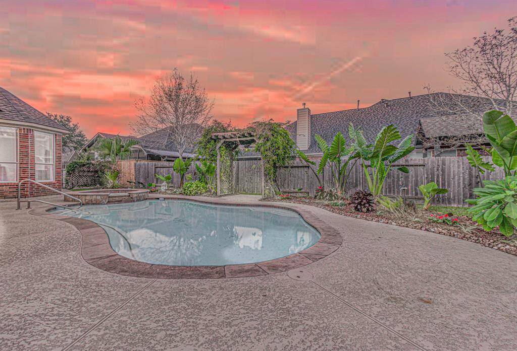 This backyard features a beautifully landscaped pool area with lush greenery and a charming pergola, set against a stunning sunset sky. Perfect for relaxation and entertaining.