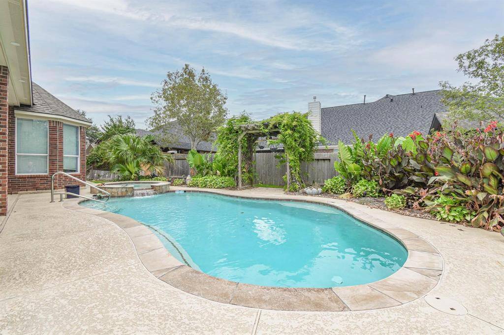 This backyard features a beautifully landscaped pool area with lush greenery and a pergola. The curved pool design is complemented by a spa section, offering a relaxing outdoor retreat.