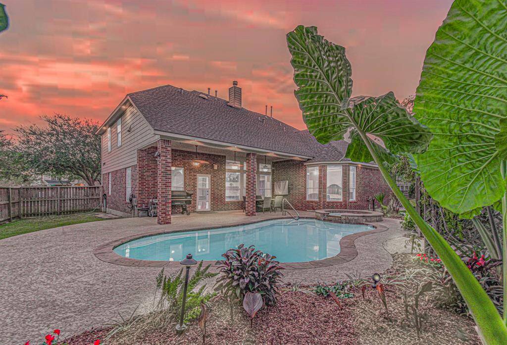This photo showcases a charming two-story brick home with a spacious backyard featuring a pool and landscaped garden. A covered patio offers a perfect spot for relaxation or entertaining, all set against a stunning sunset backdrop.