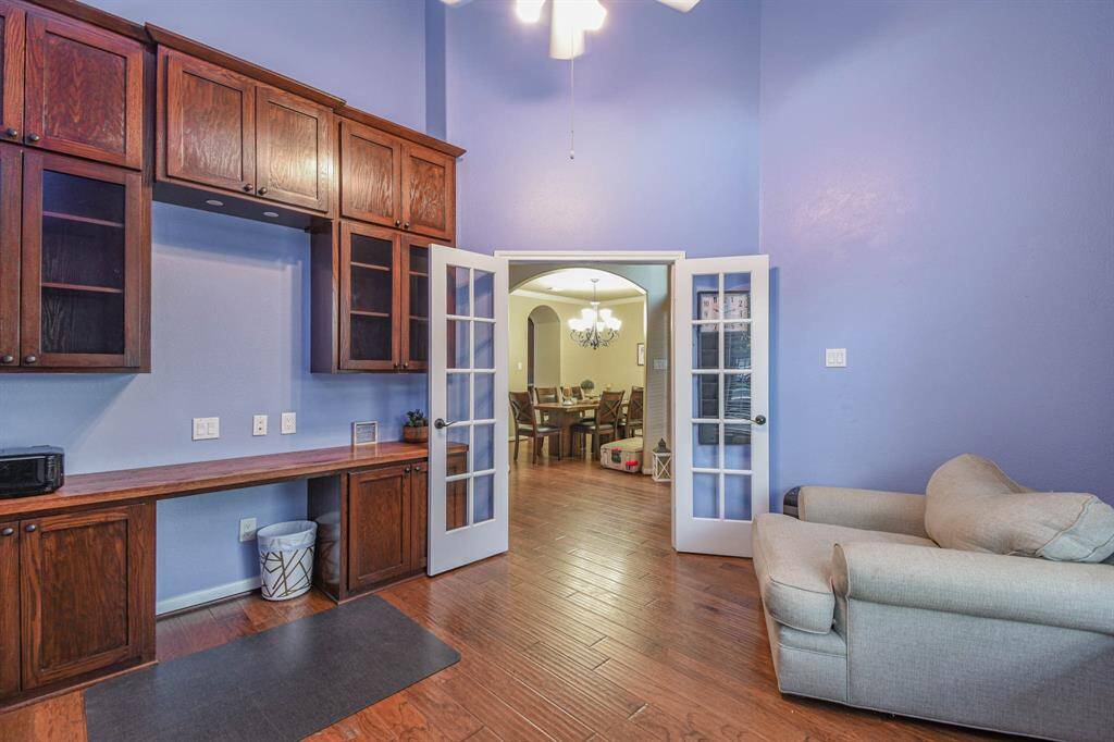 This room features rich wood cabinetry, a built-in desk, and high ceilings painted in a soothing blue. French doors open to a dining area, creating a seamless transition between spaces. The hardwood floors add warmth, and a cozy chair offers a relaxing spot.