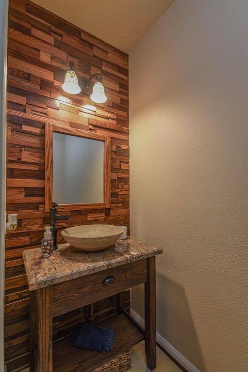 This bathroom features a stylish wooden accent wall with warm tones, a modern vessel sink on a granite countertop, and a rustic wood vanity. A wall-mounted mirror and dual light fixtures add a cozy ambiance. Perfect for a touch of elegance and warmth.