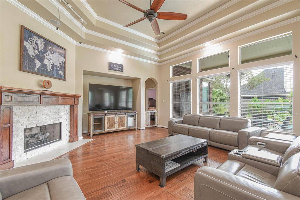 This spacious living room features high ceilings with elegant molding, a cozy fireplace, and large windows providing abundant natural light. It includes modern leather seating, a ceiling fan, and a built-in entertainment area, creating a perfect blend of comfort and style.