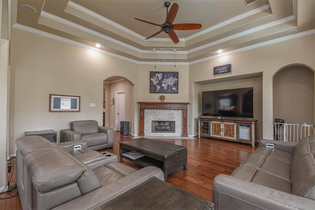 Spacious living room with elegant tray ceiling and recessed lighting. It features a cozy fireplace, large TV, and comfortable leather seating. The hardwood floors add warmth, and there's a stylish ceiling fan for comfort.