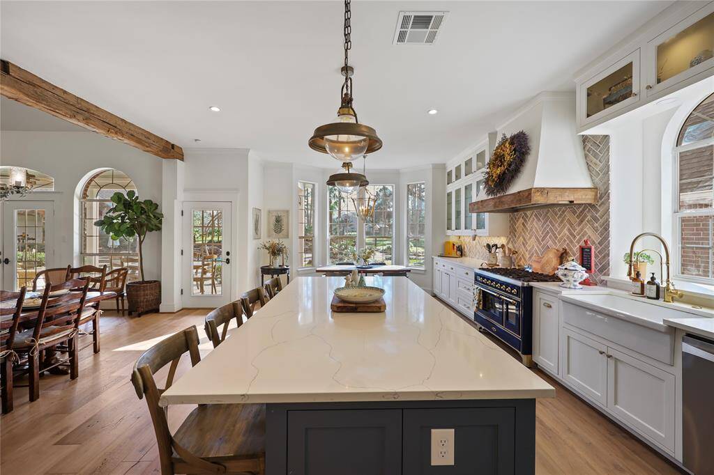 The large island with a waterfall-edge quartz countertop provides ample space for food prep and seating. The brass fixtures, including the pendant lights and faucet, complement the warm wood tones and the exposed wooden beam.