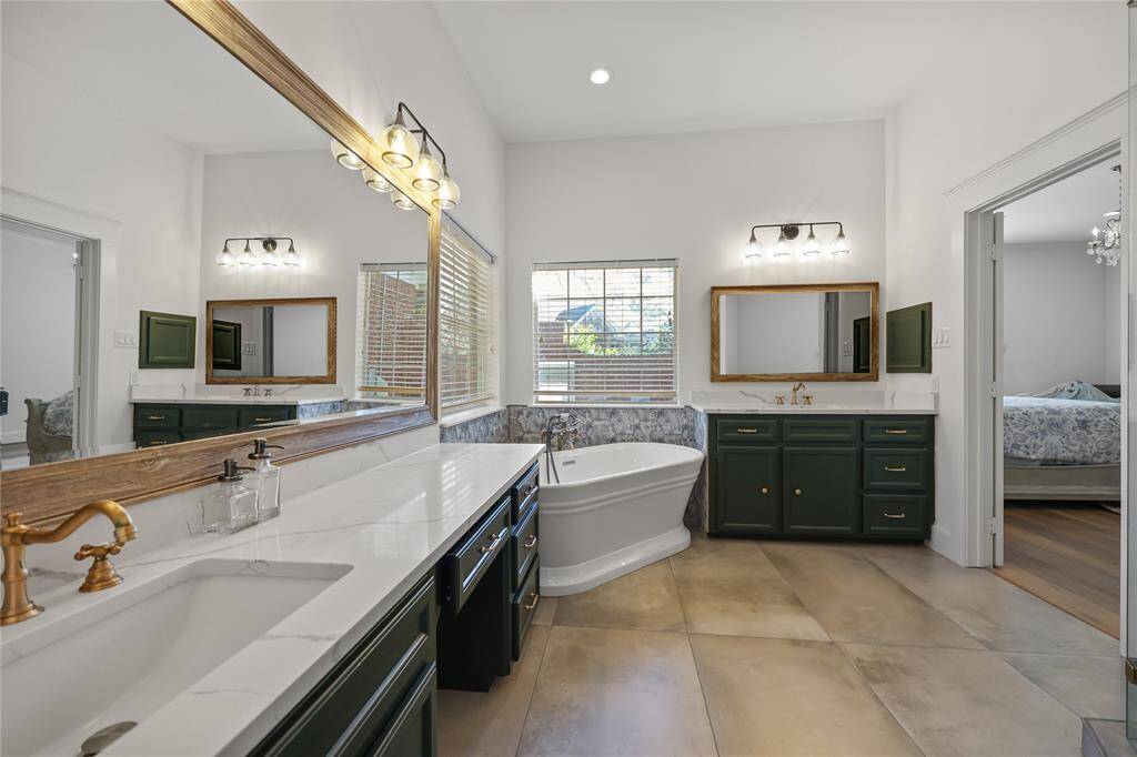 A spacious double vanity, in this recently remodeled primary ensuite bathroom, with stunning countertops, deep colored cabinetry, and gold-tone hardware. The large mirrors, framed with warm wood accents, add depth and elegance, while modern style light fixtures provide a stylish touch. A freestanding soaking tub offers a spa-like experience with natural light streams in through a generously sized window.