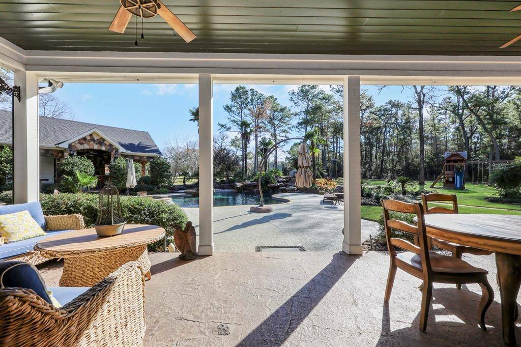 A stunning covered patio with stamped concrete overlooks a resort-style backyard with a pool, lush landscaping, and a wooded backdrop. Beyond the pool is a manicured lawn area creating a perfect setting for relaxation and outdoor entertainment.
