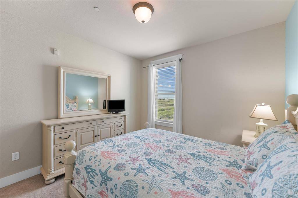 Another perspective of the guest bedroom highlights the white dresser topped with a mirror, enhancing the room's spacious feel. The coastal-themed bedding on the King-sized bed complements the light blue accent wall, creating a relaxing and inviting atmosphere.