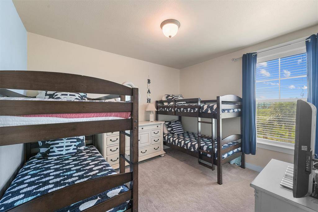 The third bedroom features two sets of wooden bunk beds with nautical-themed bedding. The room is brightly lit by a large window with blue curtains, providing a view of the outdoors. A white dresser with a lamp and beach-themed decor adds to the room's cozy ambiance.