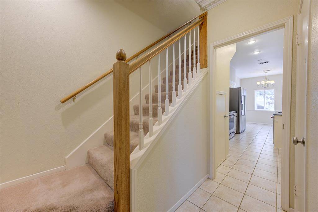 This photo showcases a well-lit hallway with tiled flooring, leading to a kitchen area. A carpeted staircase with a wooden handrail ascends to the upper level.