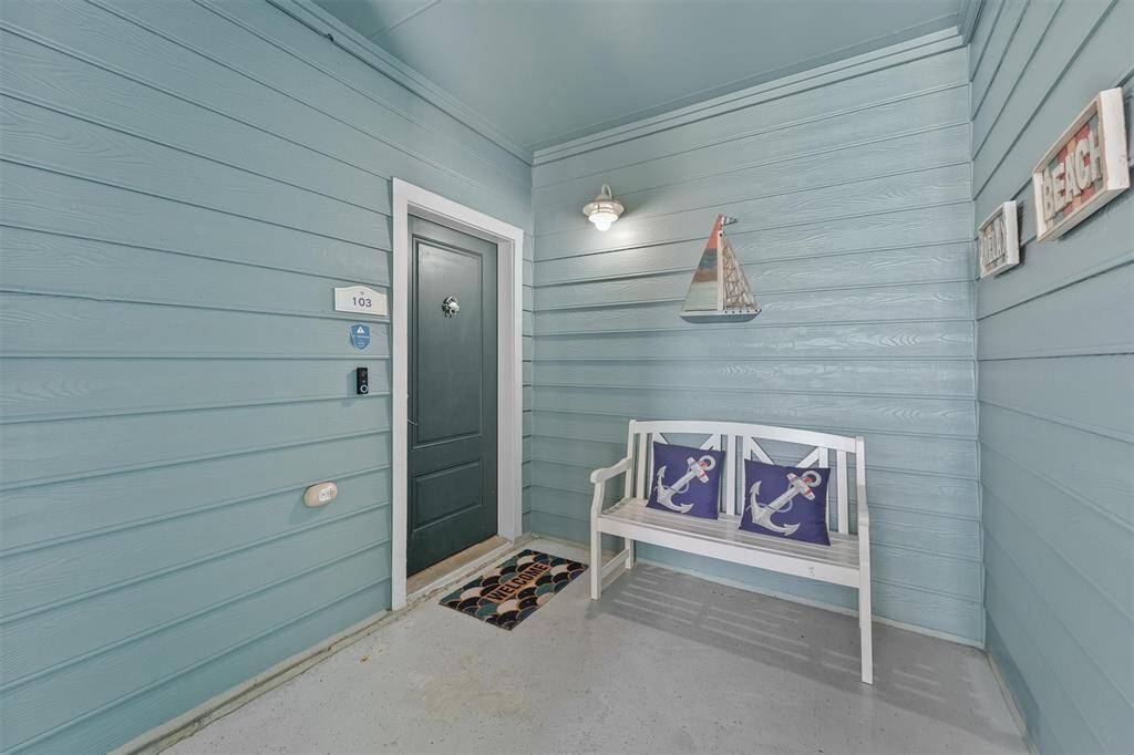 The charming entrance to condo #103 features a welcoming bench with nautical-themed pillows and coastal decor. The blue exterior and neatly arranged decorations set a pleasant tone, inviting guests to enjoy a relaxing seaside retreat.