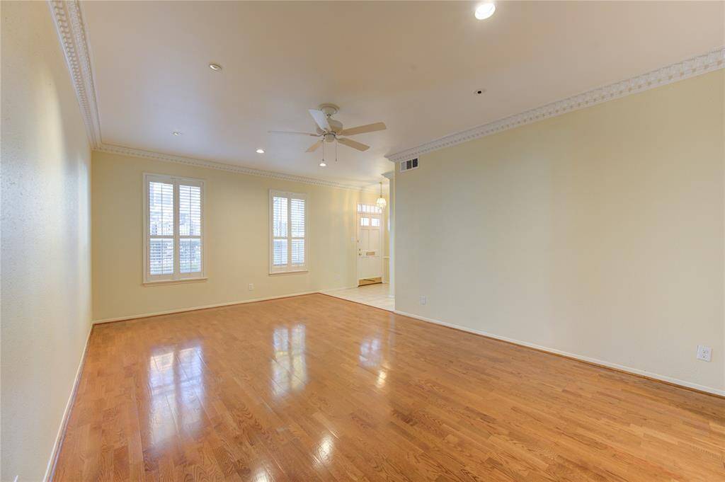 This spacious living room features engineered wood floors, neutral walls, and crown molding. It has recessed lighting, a ceiling fan, and large windows with shutters for natural light.