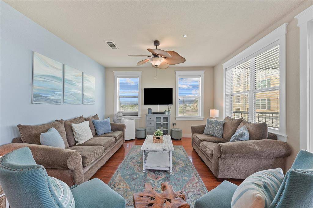 This living room boasts abundant natural light from large windows, creating a bright and inviting space. The room features a ceiling fan with leaf-shaped blades and serene ocean-themed wall art, enhancing the coastal ambiance.