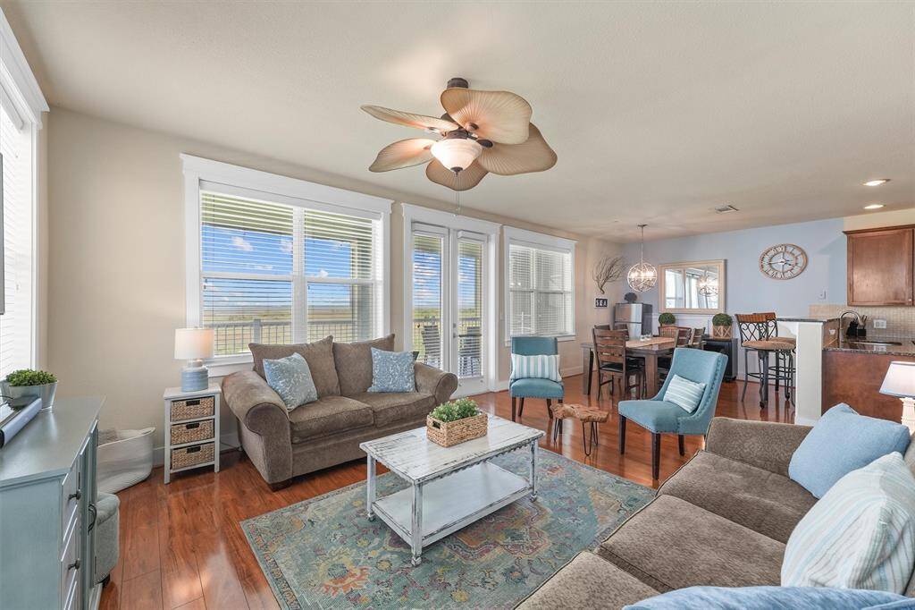 This view of the living room shows its open layout, seamlessly connecting to the dining area and kitchen. The large windows and French doors flood the space with natural light, while the decor maintains a cohesive coastal theme, perfect for a bright and airy atmosphere.