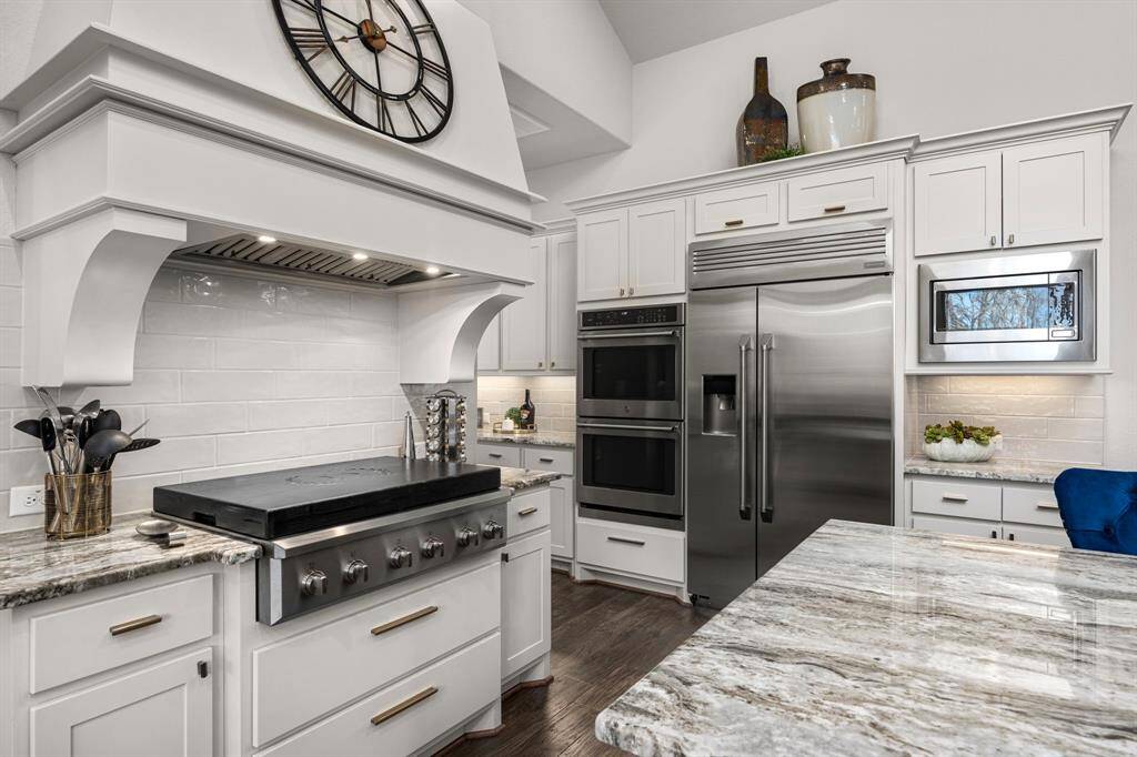 This modern kitchen features sleek stainless steel appliances, including a double oven and large fridge. It has elegant white cabinetry, a spacious marble countertop, and a stylish range hood with a decorative clock. The space is bright and functional, perfect for cooking and entertaining.