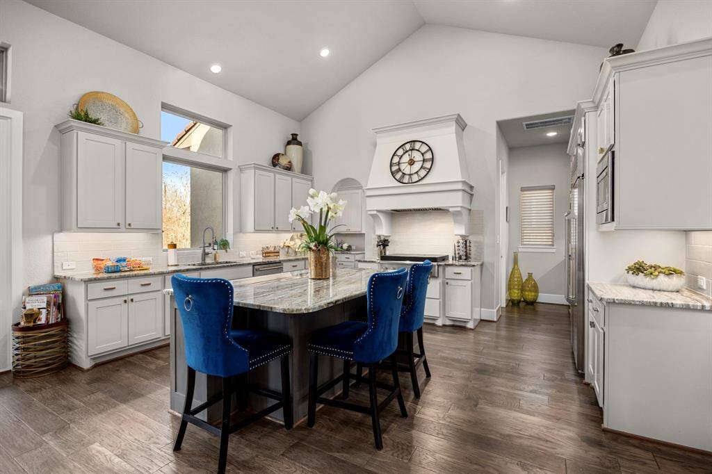 This kitchen features high ceilings and an open layout with elegant white cabinetry and a large island. It includes modern appliances, a stylish range hood with a clock, and beautiful wood flooring. Bright and welcoming, perfect for entertaining.