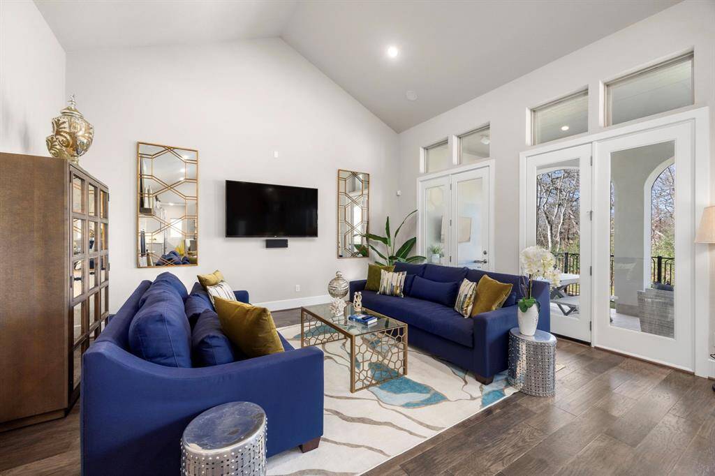 This living room features high ceilings and abundant natural light, with large windows and French doors leading to an outdoor space. It has elegant decor, including blue sofas, a modern coffee table, and stylish mirrors. The open layout and hardwood floors create a spacious and inviting atmosphere.